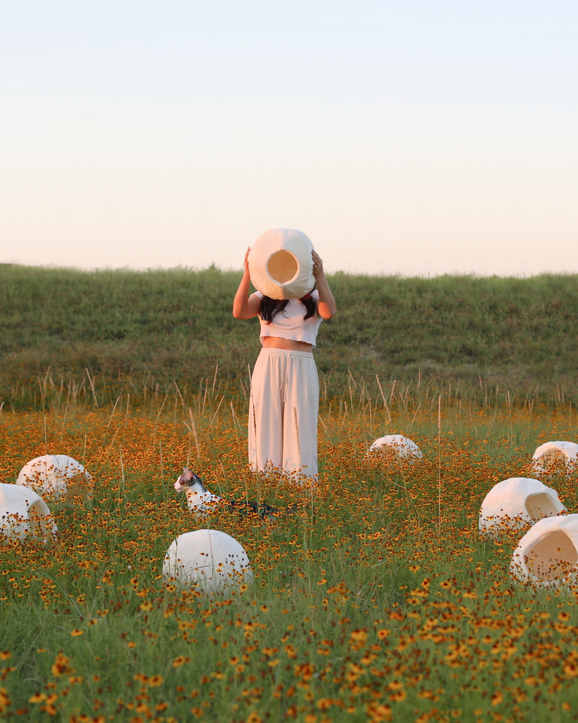 Wicksie Tu, founder of Catenary Home, standing in a field of flowers holding a modern cat cave bed above her head, surrounded by multiple cat caves and a curious cat. Showcasing Catenary Home's best cat cave design, merging nature with contemporary cat furniture.