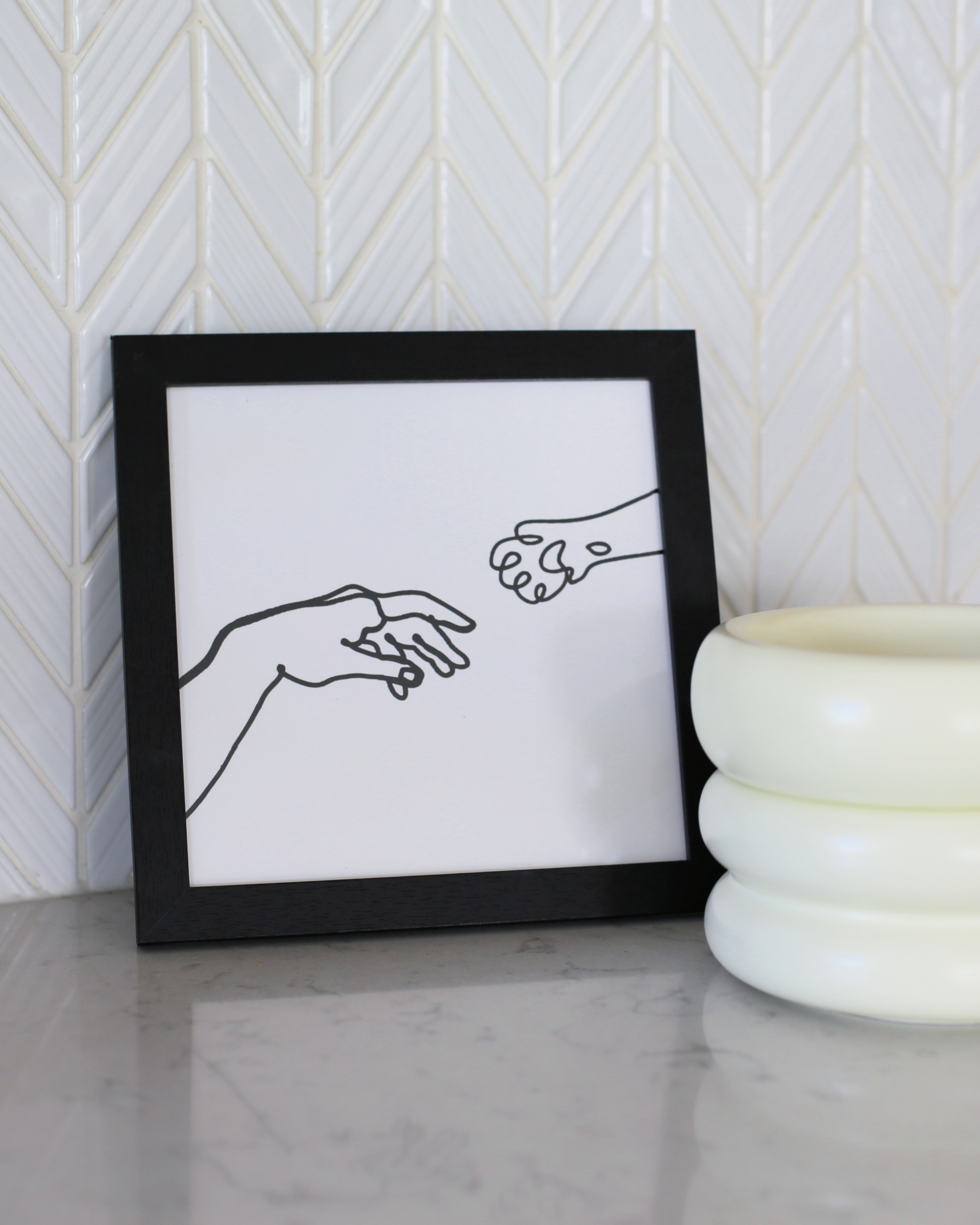 single line art of hand and paw reaching in black frame by catenary home modern cat furniture company  on marble counter with white backsplash and elevated cat bowls next to it