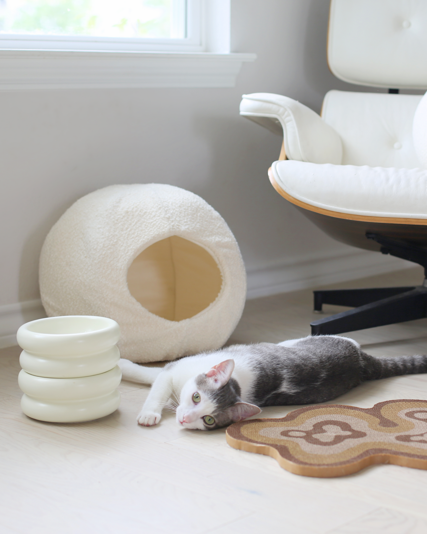 grey and white aesthetic cat laying next to boucle cat cave bed and the best elevated cat bowls with modern cat scratcher and mid century modern eames loung chair in background by catenary home modern cat furniture company